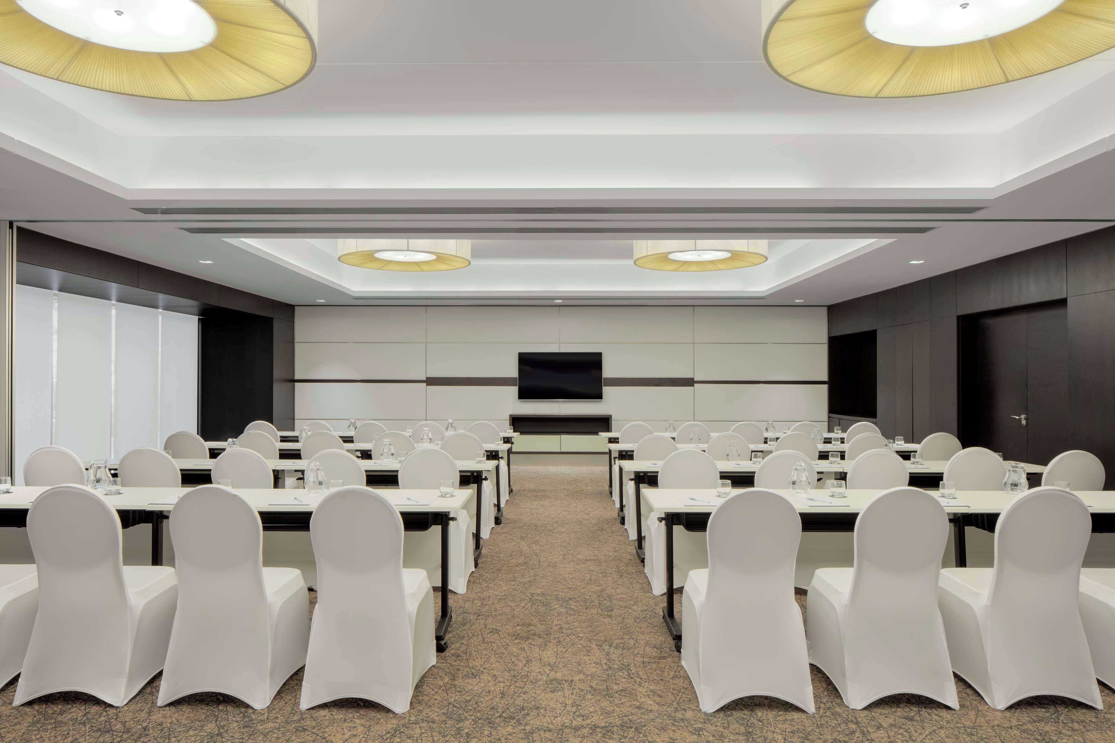 A conference room with rows of tables and chairs, a large screen, and ceiling lights.