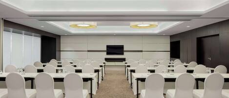 Un salón de conferencias con filas de mesas y sillas, una pantalla grande y luces en el techo.
