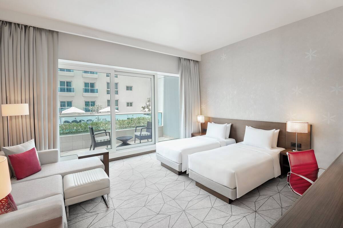 A hotel room with two beds, a sofa, a chair, and a balcony with a view of buildings and greenery.
