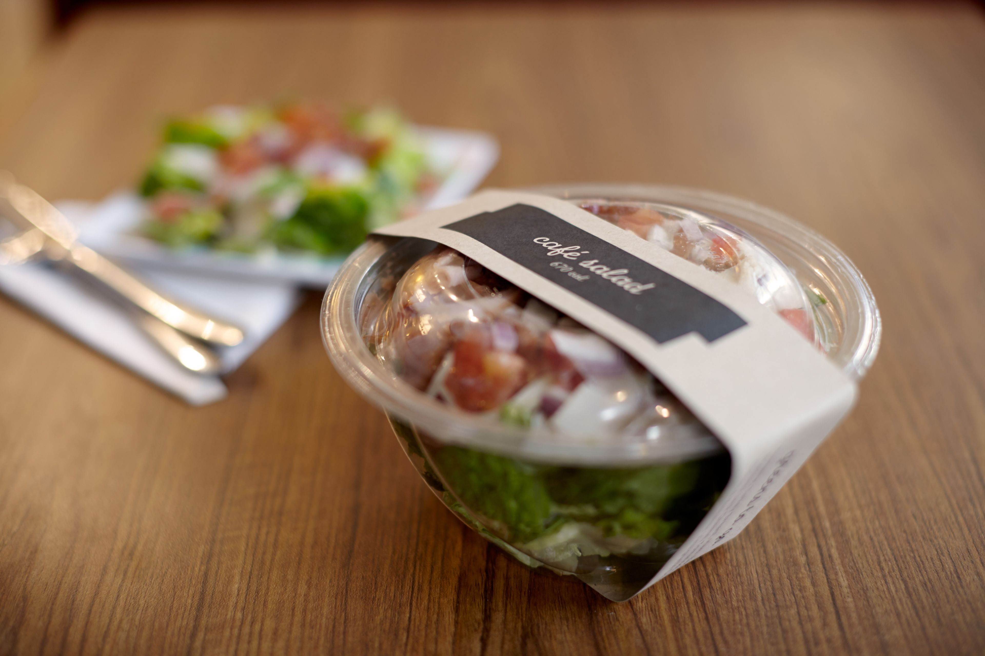 Une salade dans un récipient en plastique transparent, muni d’une étiquette, posée sur une table en bois.