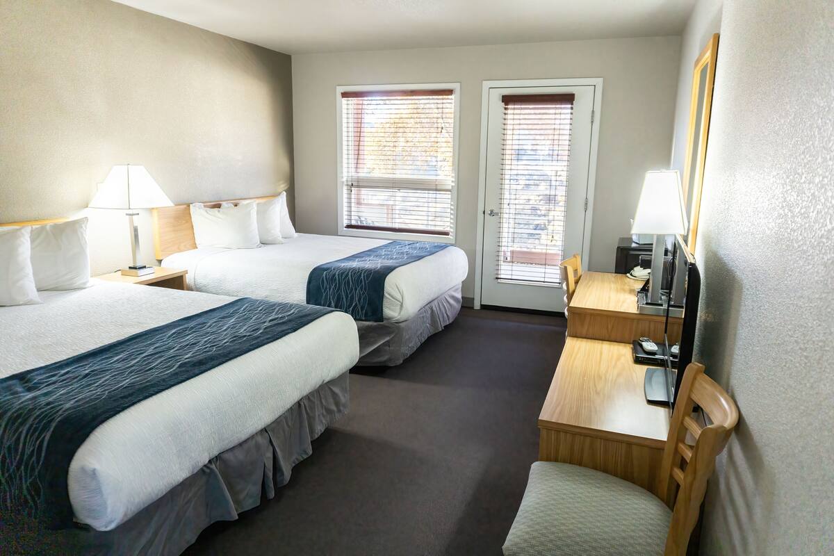 Quarto de hotel com duas camas, uma mesa, uma cadeira, uma televisão e uma janela com persianas.