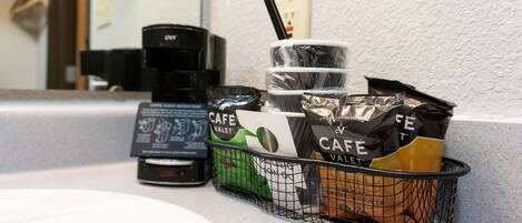 Una cafetera, una taza de café y una cesta con paquetes de café sobre una encimera.