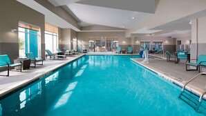 Una piscina interior con agua cristalina, rodeada de sillas de descanso y un panel de vidrio.