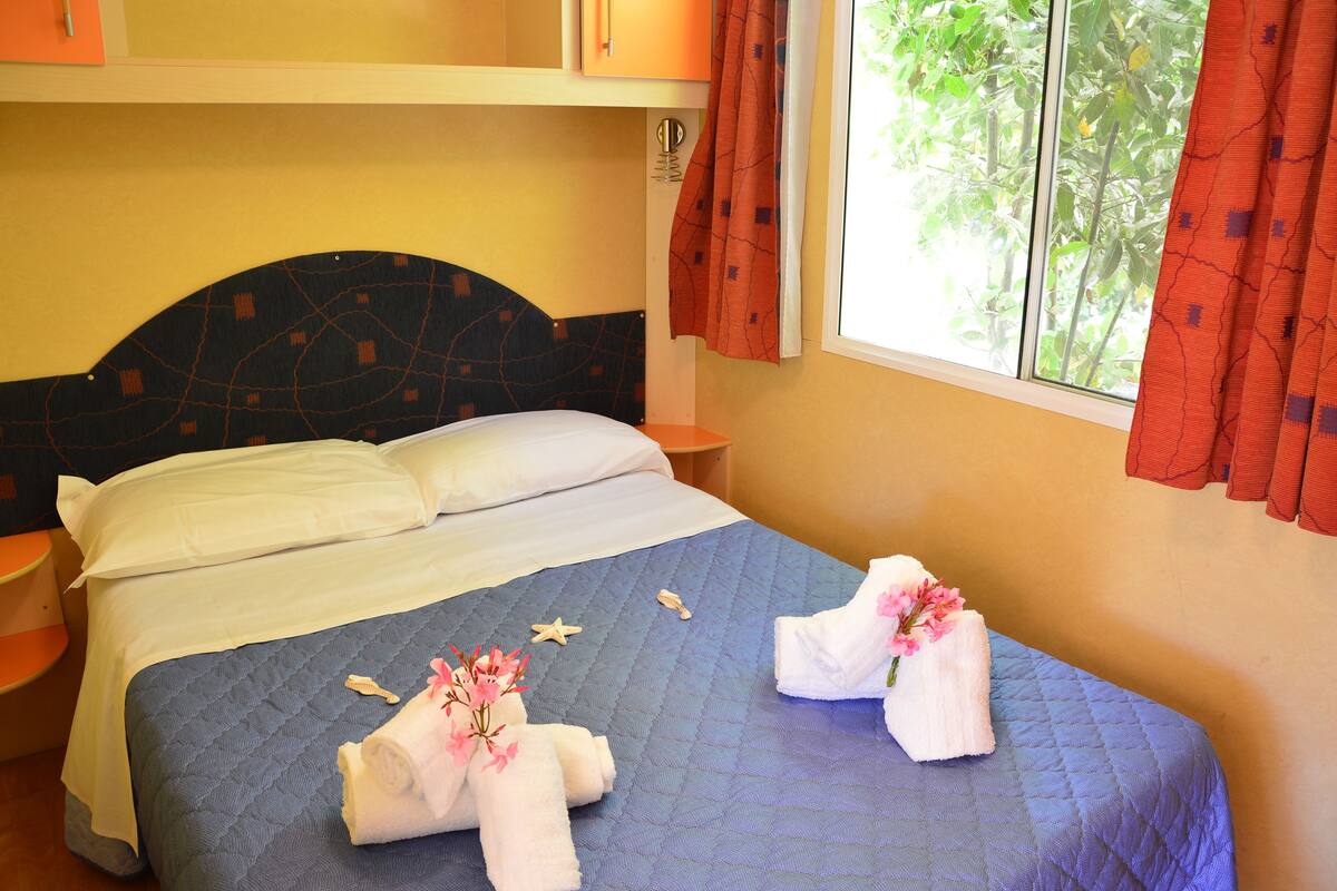 A neatly made bed with blue bedding, white pillows, and two towels with pink flowers.
