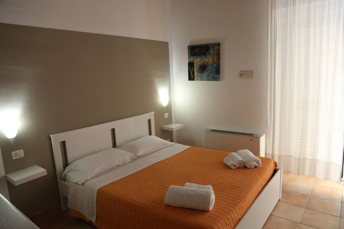 A hotel room with a bed, white pillows, an orange bedspread, two white towels, a nightstand, a small table, a wall-mounted artwork, and a window with curtains.