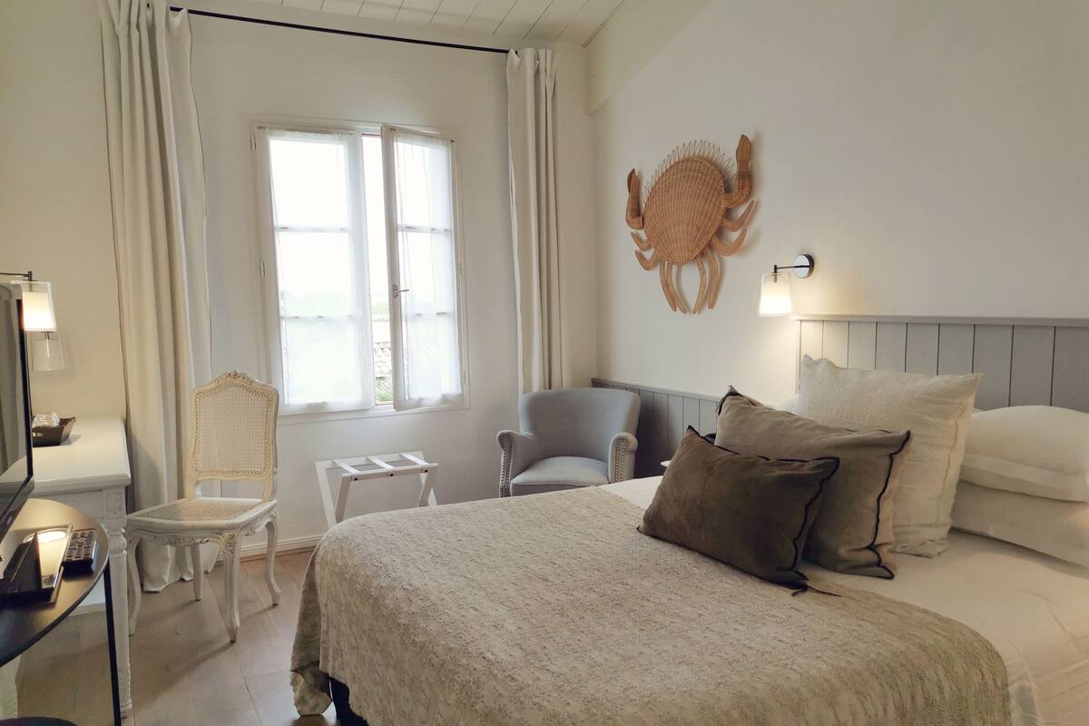 A hotel room with a large bed, two chairs, a small table, a wall-mounted lamp, and a window with curtains.