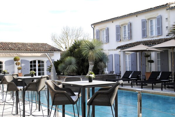 A poolside area with wicker chairs and tables, a pool, and a building with shutters. - Le Clos Saint-Martin Hôtel & Spa (Saint-Martin-de-Re)