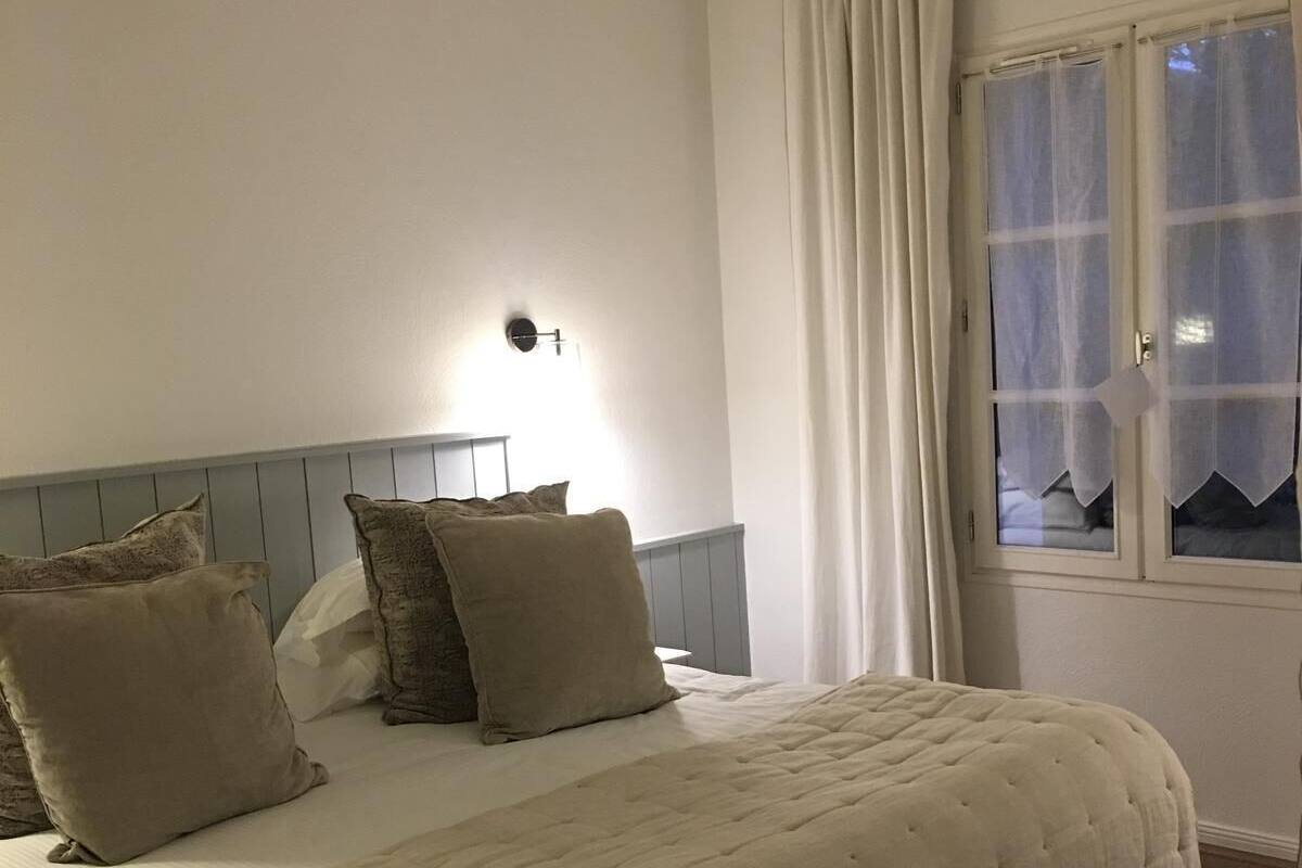 A bedroom with a bed, pillows, a nightstand, a wall-mounted lamp, and a window with curtains.