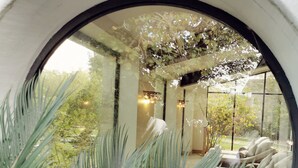 Finestra rodona amb vista a zona d'estar interior, amb un sofà i una taula, envoltada de vegetació.