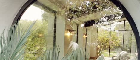 Una ventana circular con vistas a un área de estar interior, que incluye un sofá y una mesa, rodeada de follaje.