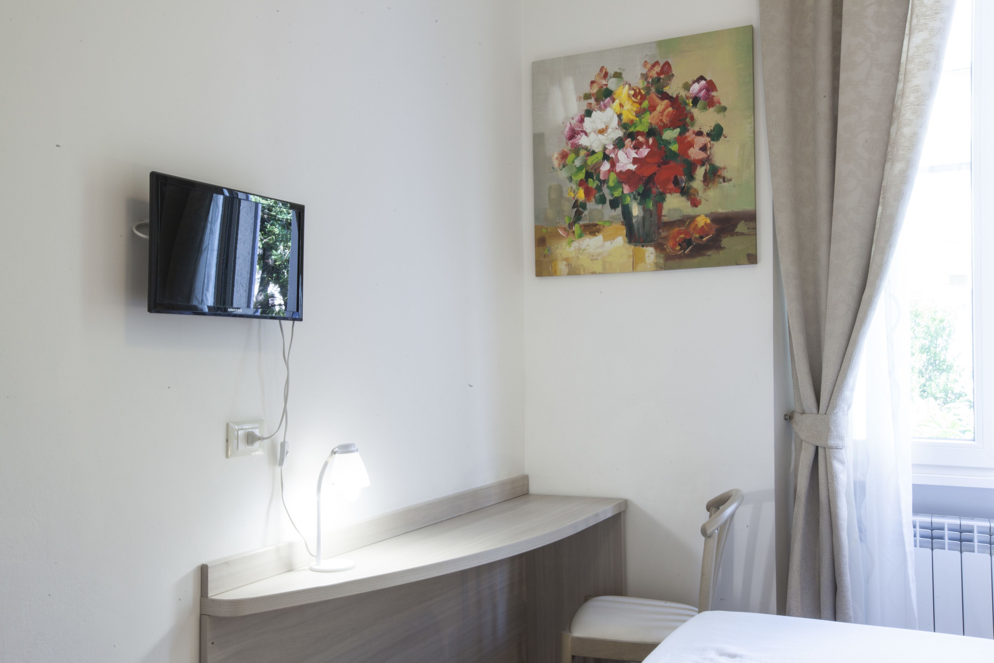A hotel room with a flat-screen TV mounted on the wall, a bedside table with a lamp, a chair, and a painting of flowers on the wall.