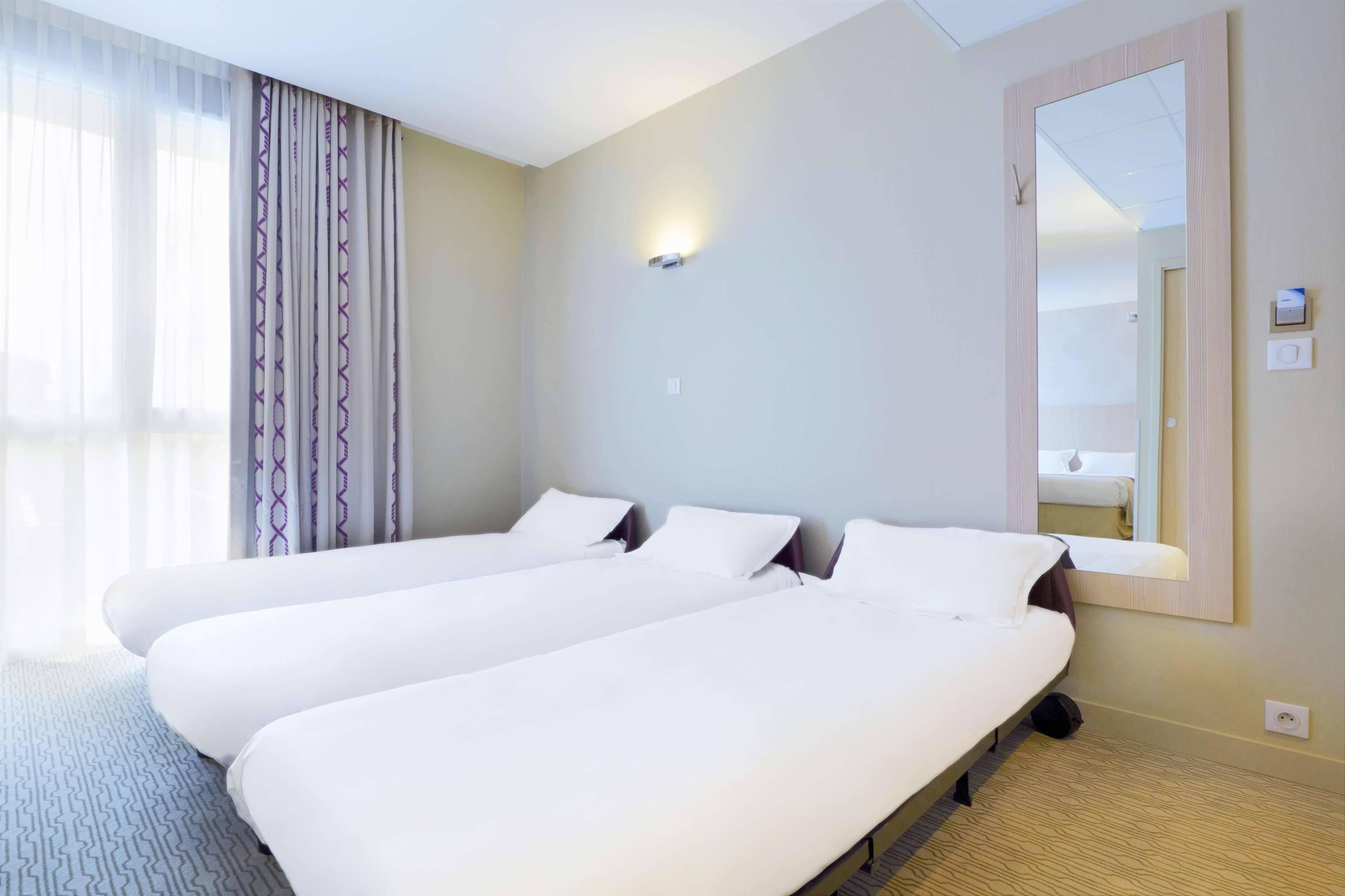 A hotel room with two single beds, a large mirror, and a window with sheer curtains.