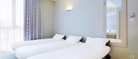 Una habitación de hotel con dos camas individuales, un espejo grande y una ventana con cortinas translúcidas.
