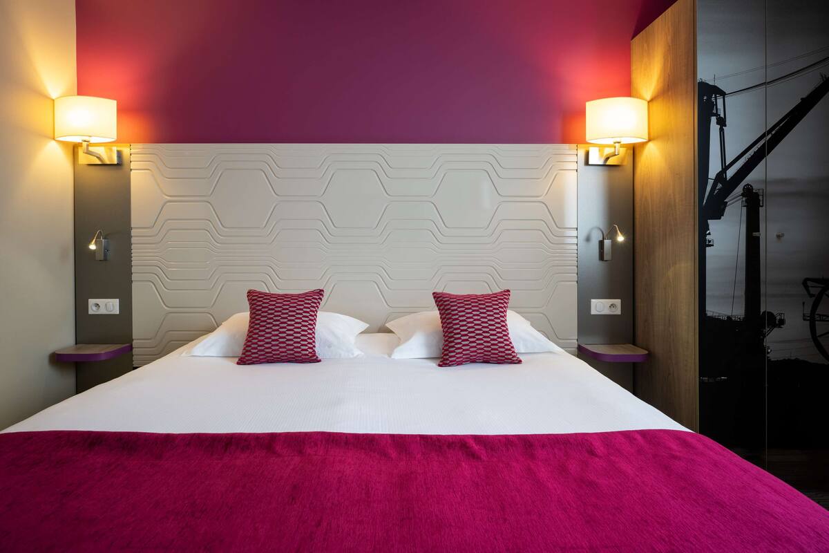 Una habitación de hotel con una cama grande, dos almohadones rojos estampados, un cabecero blanco y dos lámparas fijadas a la pared.