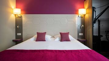 Une chambre d’hôtel avec un grand lit, deux coussins rouges à motifs, une tête de lit blanche et deux lampes fixées au mur.