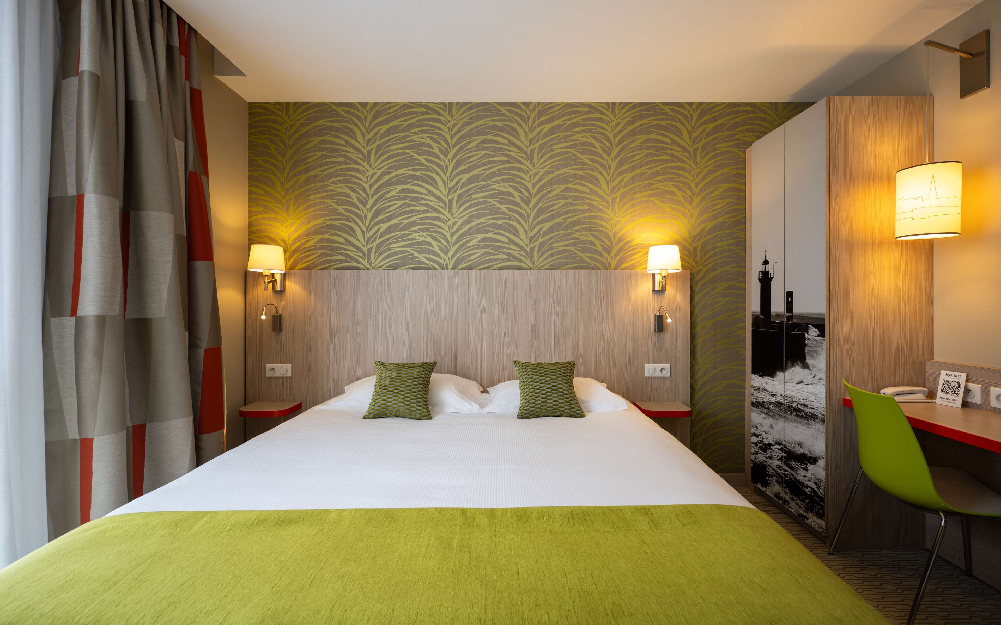 Une chambre d’hôtel avec un grand lit, deux lampes de chevet, un bureau avec une chaise et un mur orné d’une image de phare.
