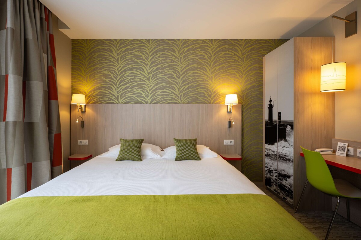 Une chambre d’hôtel avec un grand lit, deux lampes de chevet, un bureau avec une chaise et un mur orné d’une image de phare.