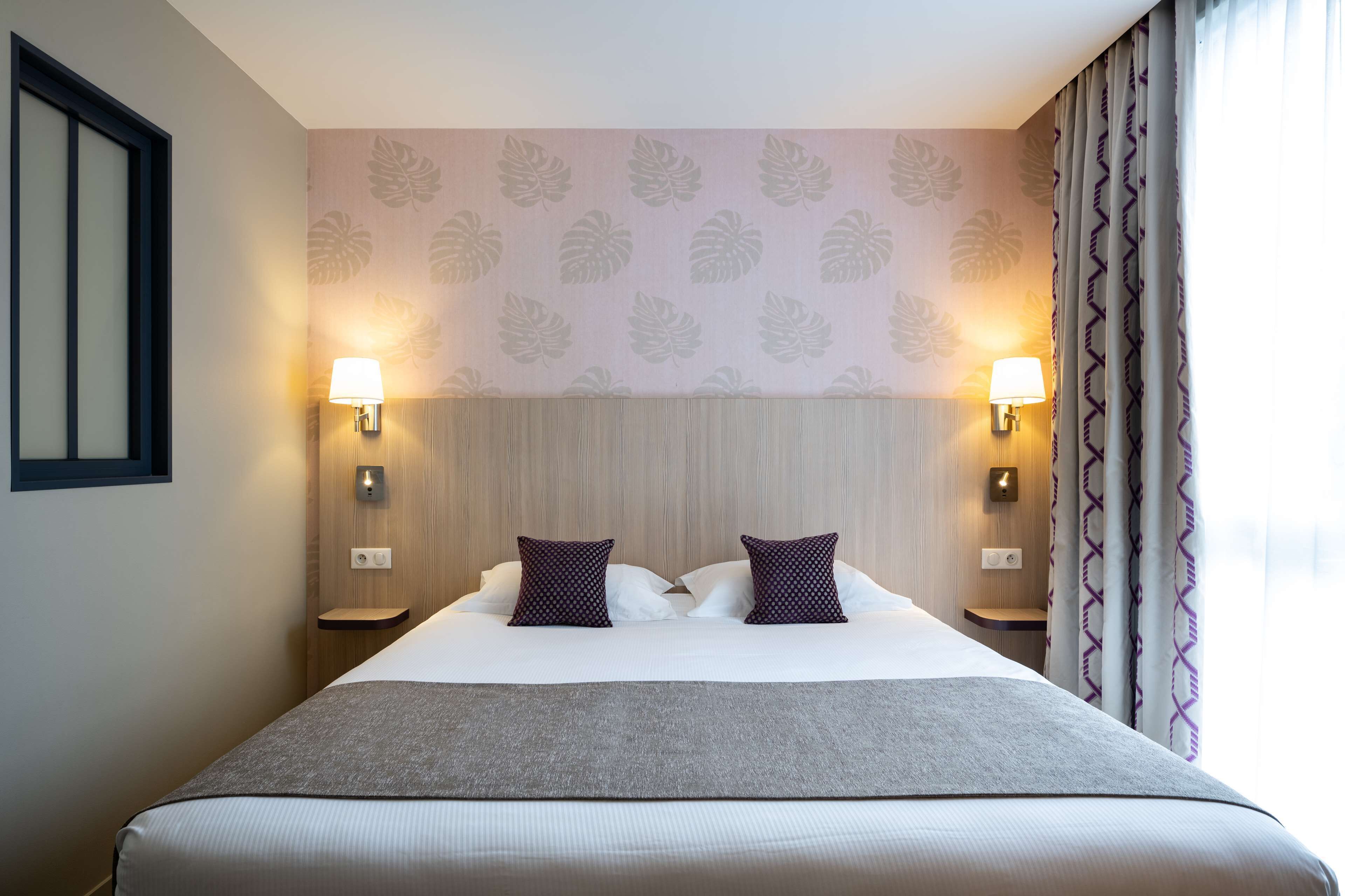 Habitación de hotel con una cama grande, dos lámparas fijadas a la pared, un cabecero de madera y un papel tapiz estampado.