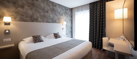 A hotel room with a large bed, a desk, and a mirror. There are wall-mounted lights and a window with curtains.