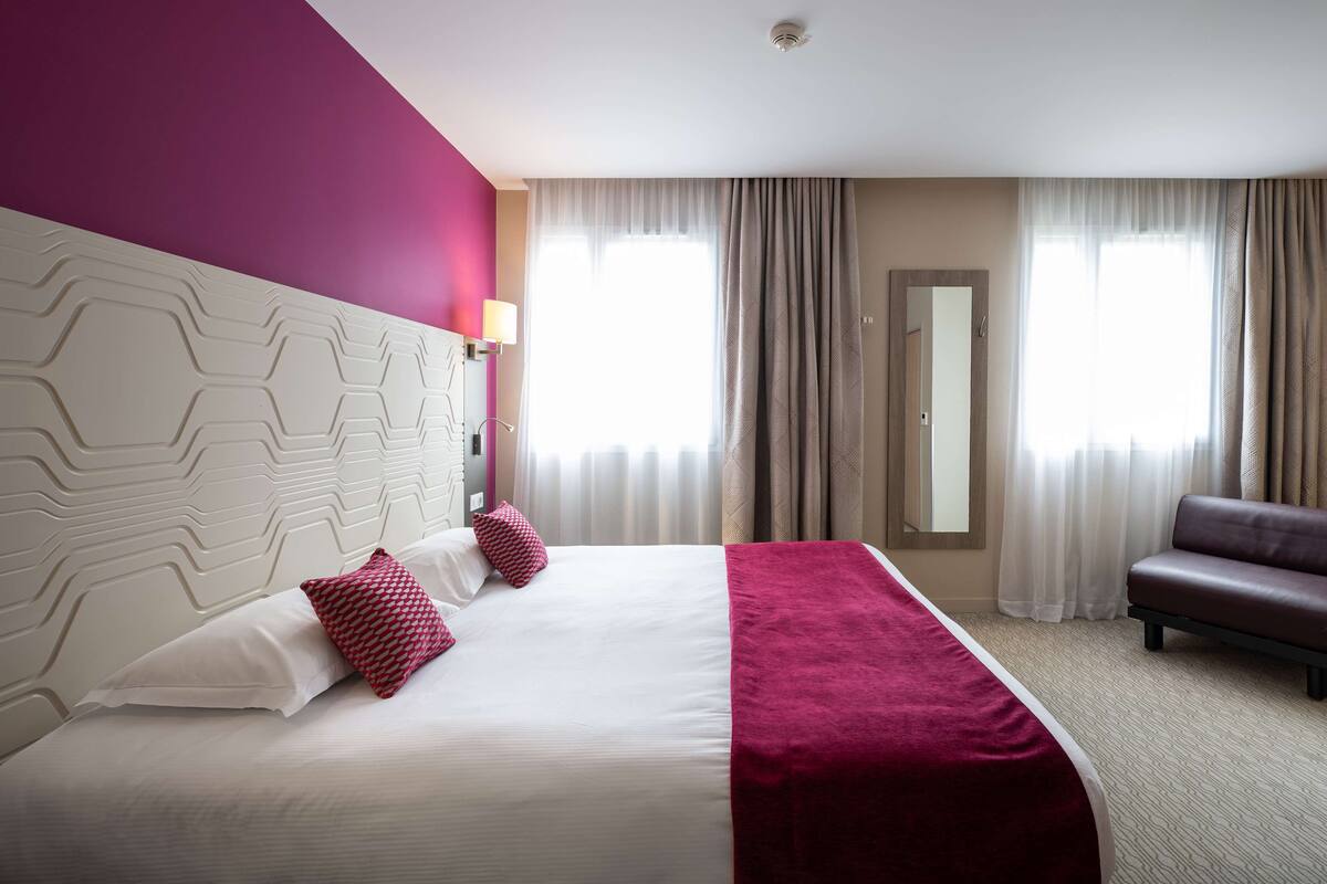 Una habitación de hotel con una cama grande, un cabecero de estampado geométrico, dos almohadas rojas, un banco de cuero marrón y un espejo en la pared.