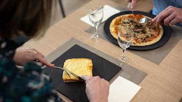 A dining table with a pizza and a slice of bread on a black plate, a wine glass, and a person cutting the bread.