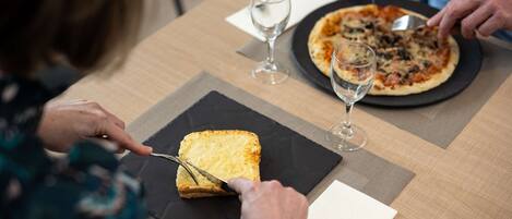 A dining table with a pizza and a slice of bread on a black plate, a wine glass, and a person cutting the bread.