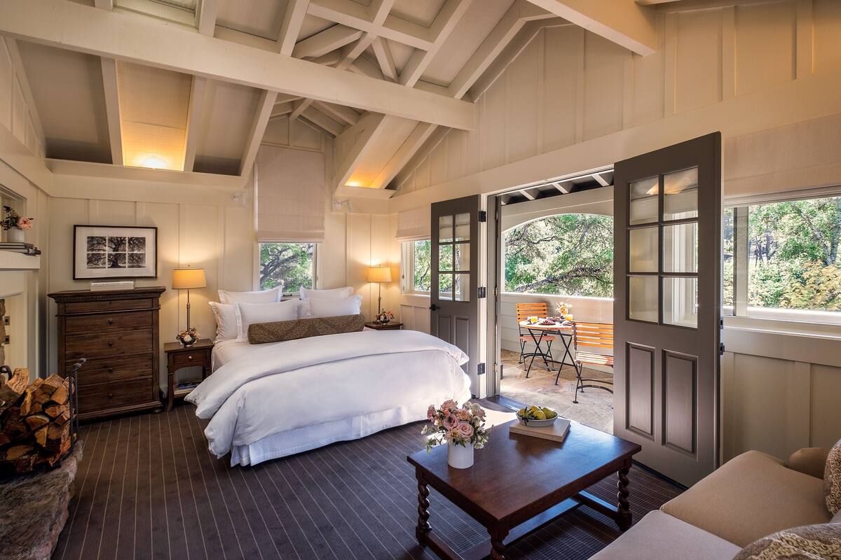 A bedroom with a large bed, a dresser, a nightstand with a lamp, a wooden coffee table, and a view of the outdoors through an open door.
