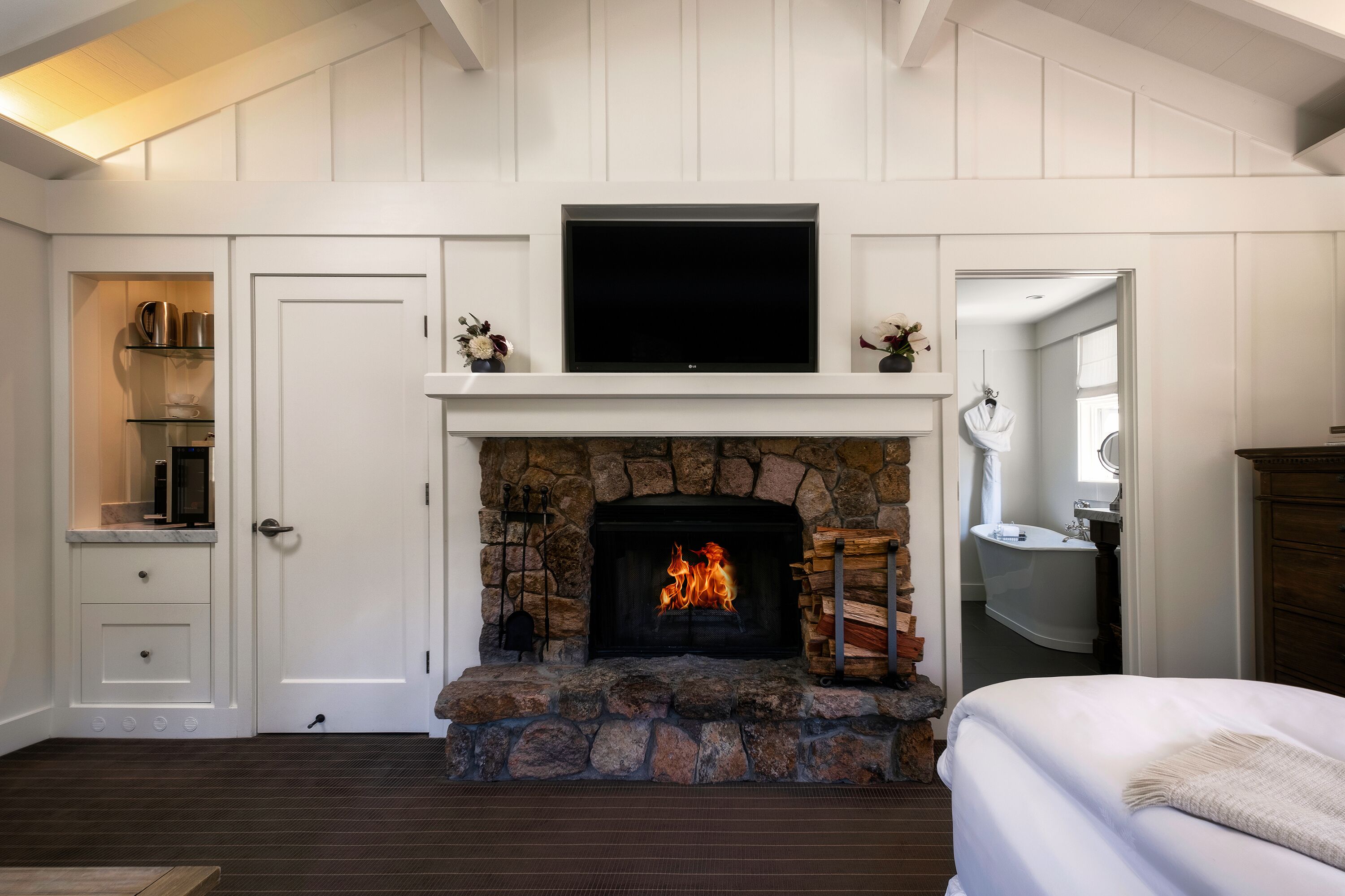A room with a stone fireplace, a television above it, and a bed with a white blanket.