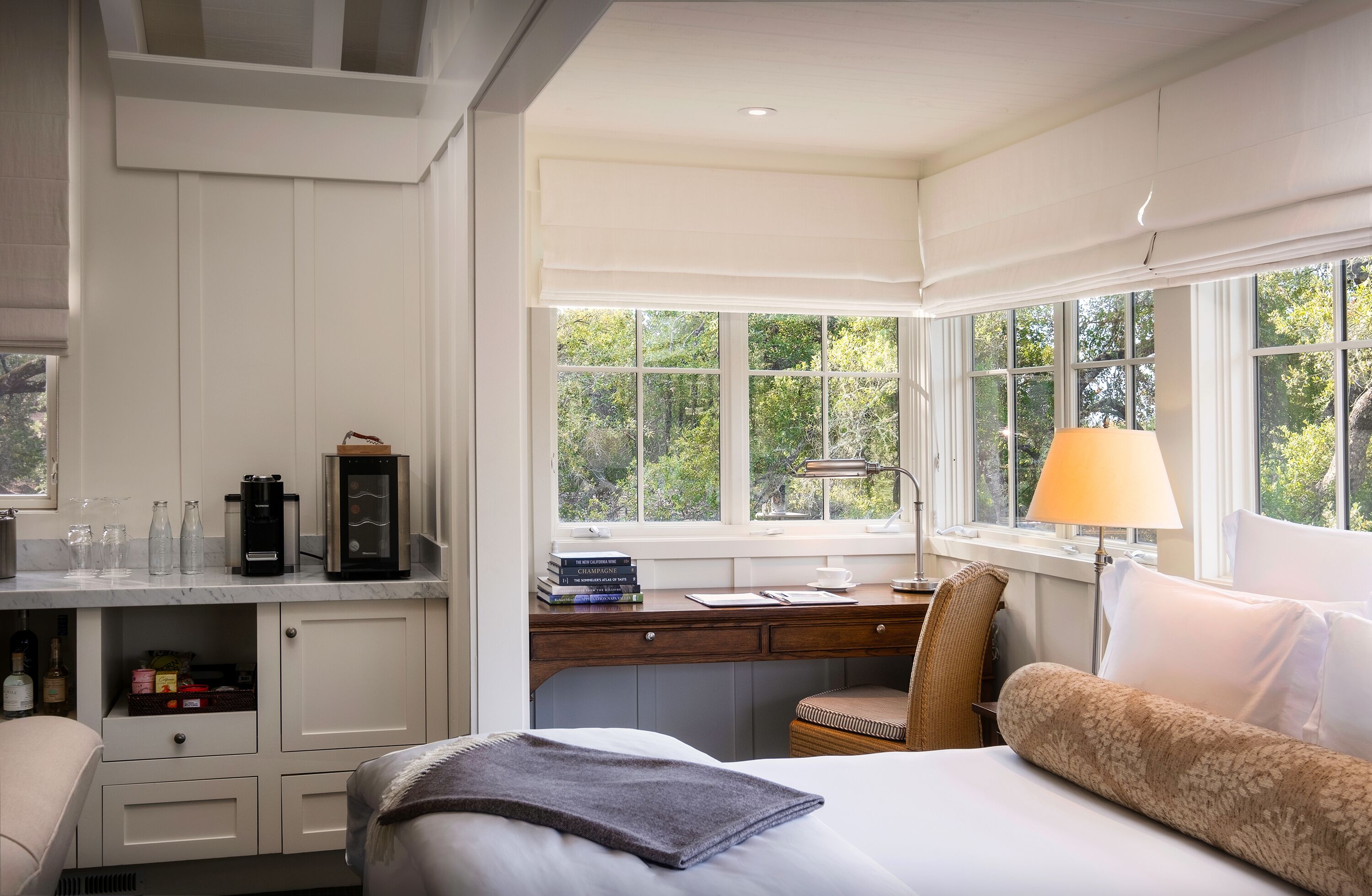 Habitación de hotel con cama, escritorio con lámpara, ventana con vistas a árboles y un minibar.