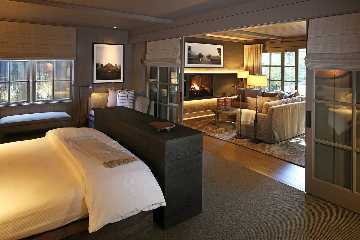 A modern hotel room with a large bed, a seating area with a sofa and armchairs, a fireplace, and a view through a glass door.