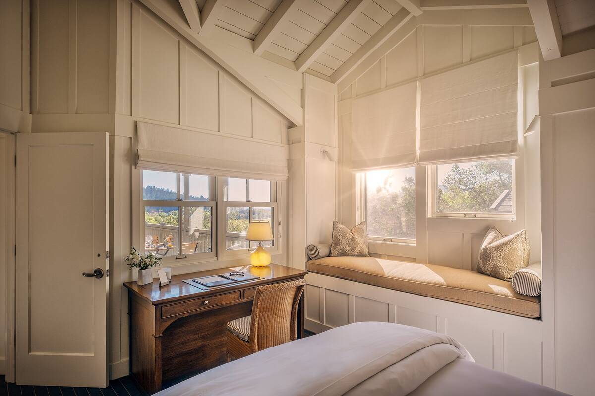 A bedroom with a desk, chair, and a window seat with cushions.