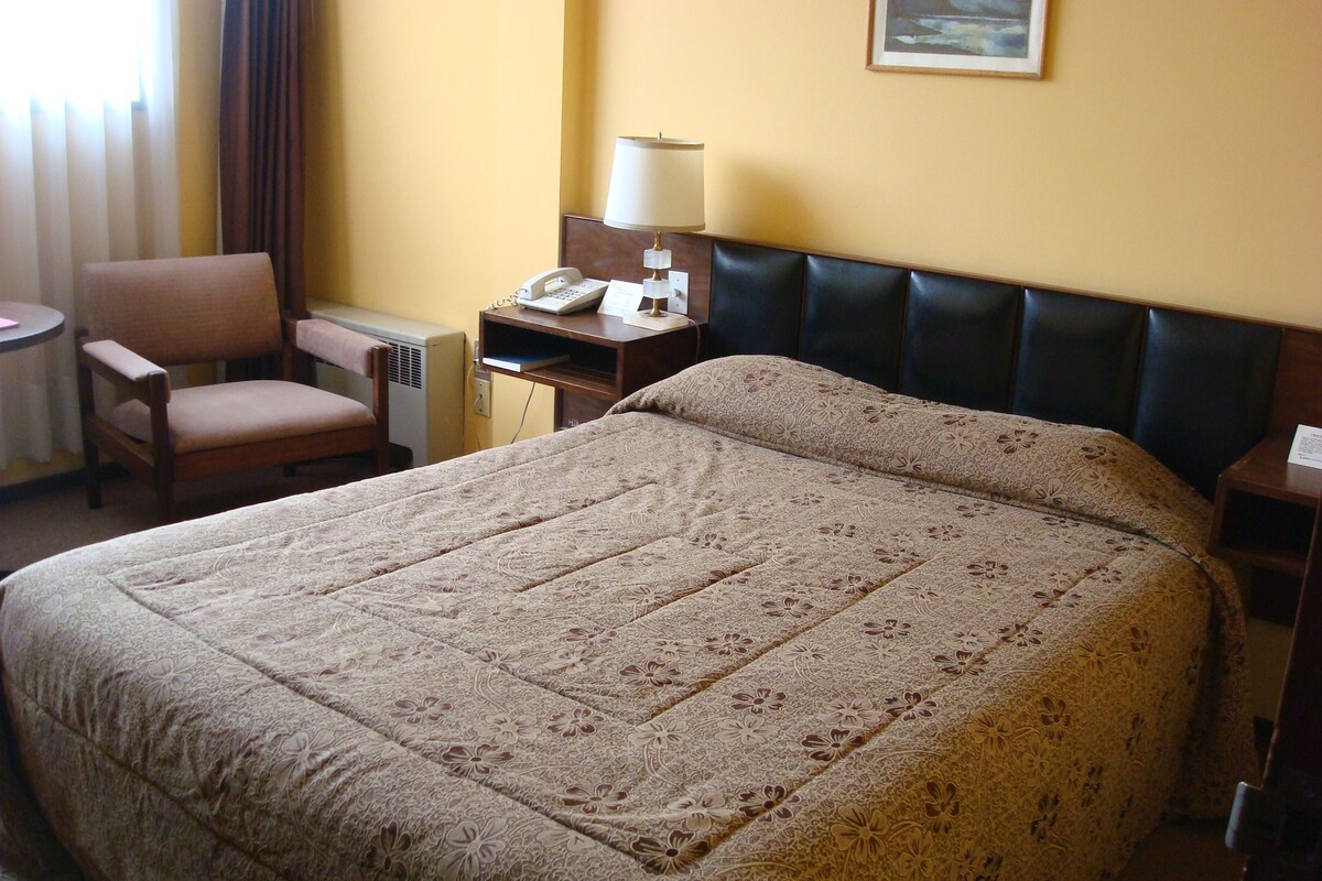 A hotel room with a large bed, a chair, a desk with a lamp, and a painting on the wall.