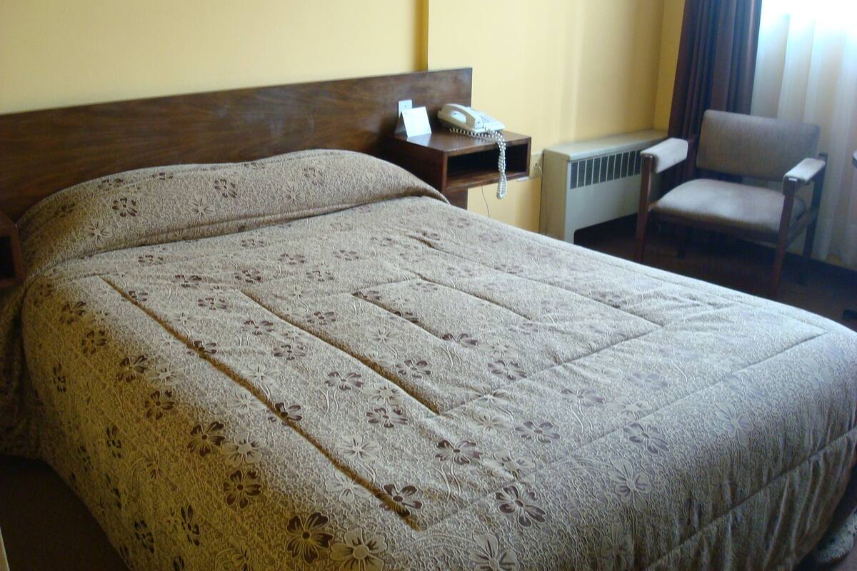 A hotel room with a large bed, a bedside table with a telephone, an air conditioning unit, and a chair.