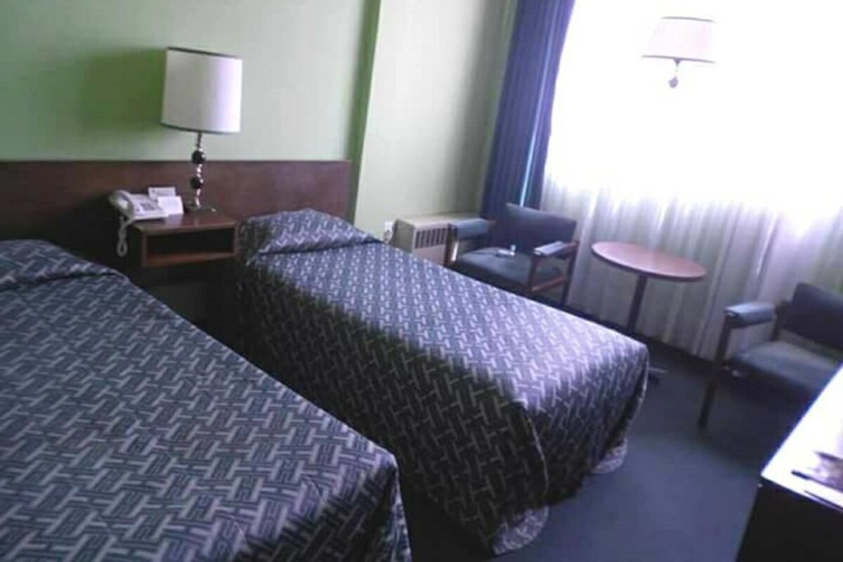 A hotel room with two beds, a desk with a lamp, a chair, a small table, and a window with curtains.