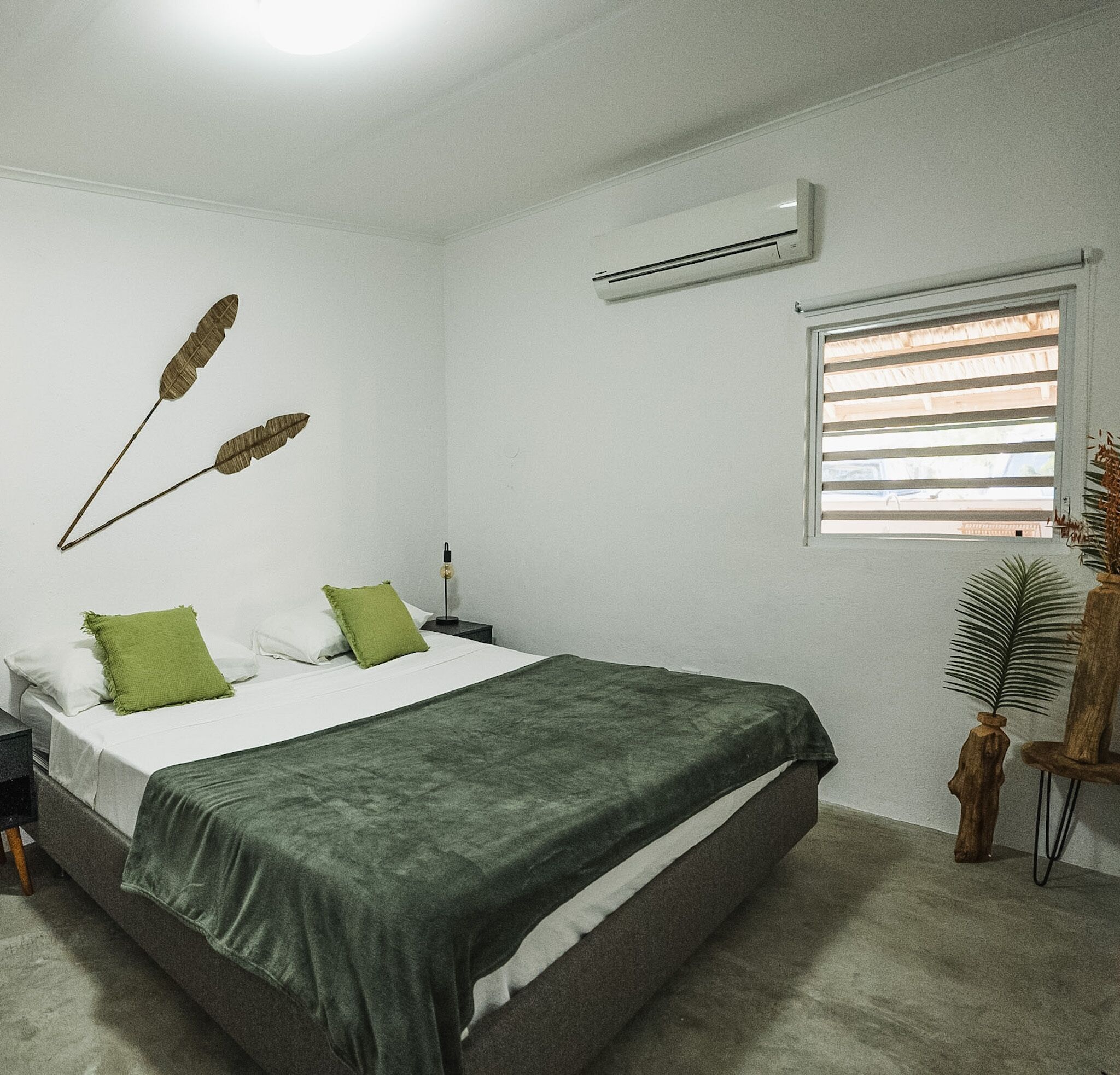 A bedroom with a bed, pillows, an air conditioner, a window with blinds, and a decorative plant.