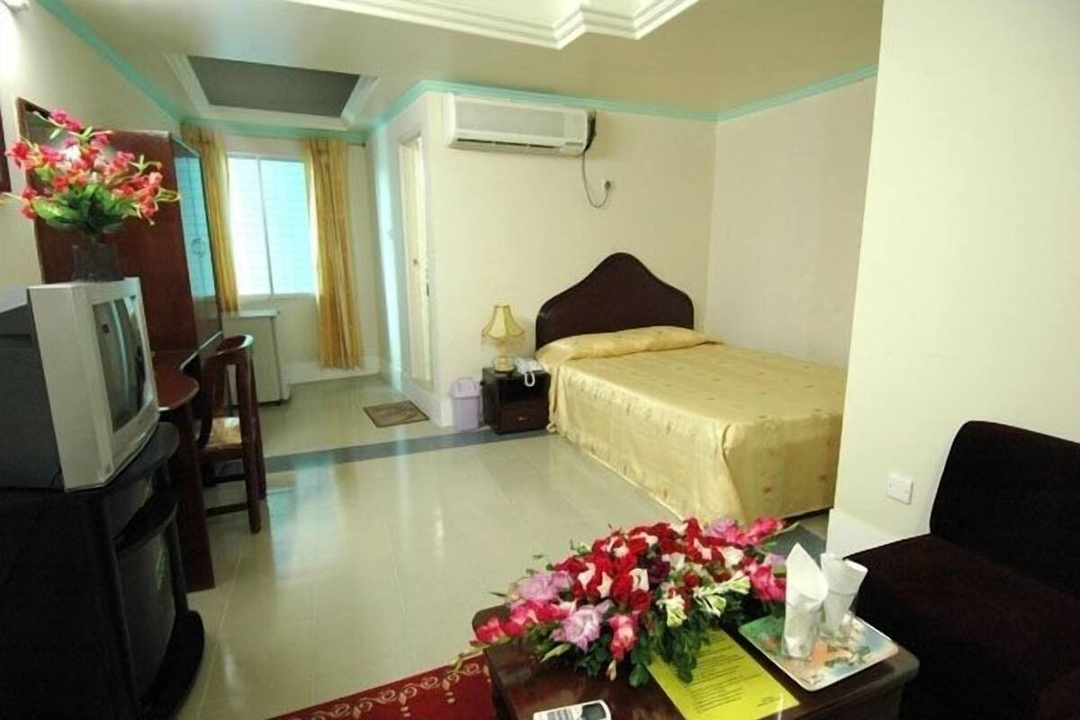 A hotel room with a bed, a TV, a desk with flowers, and a window with curtains.