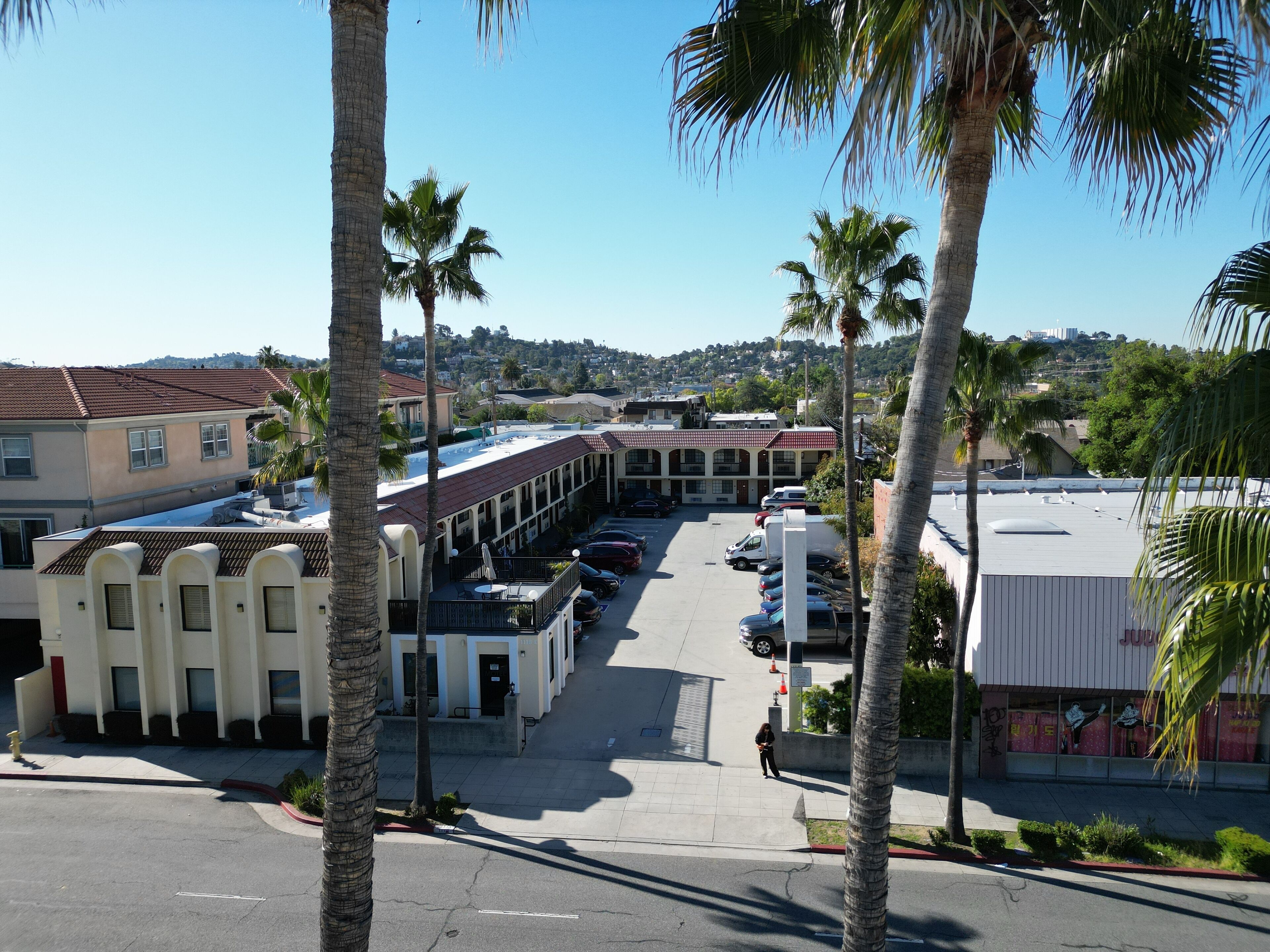 A parking lot with several cars, a building with a red roof, and palm trees.