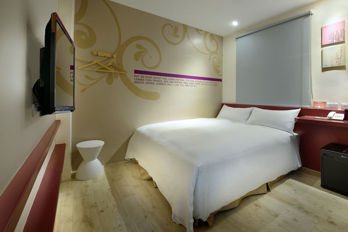 A hotel room with a bed, a TV mounted on the wall, a small white pedestal, and a wooden floor.