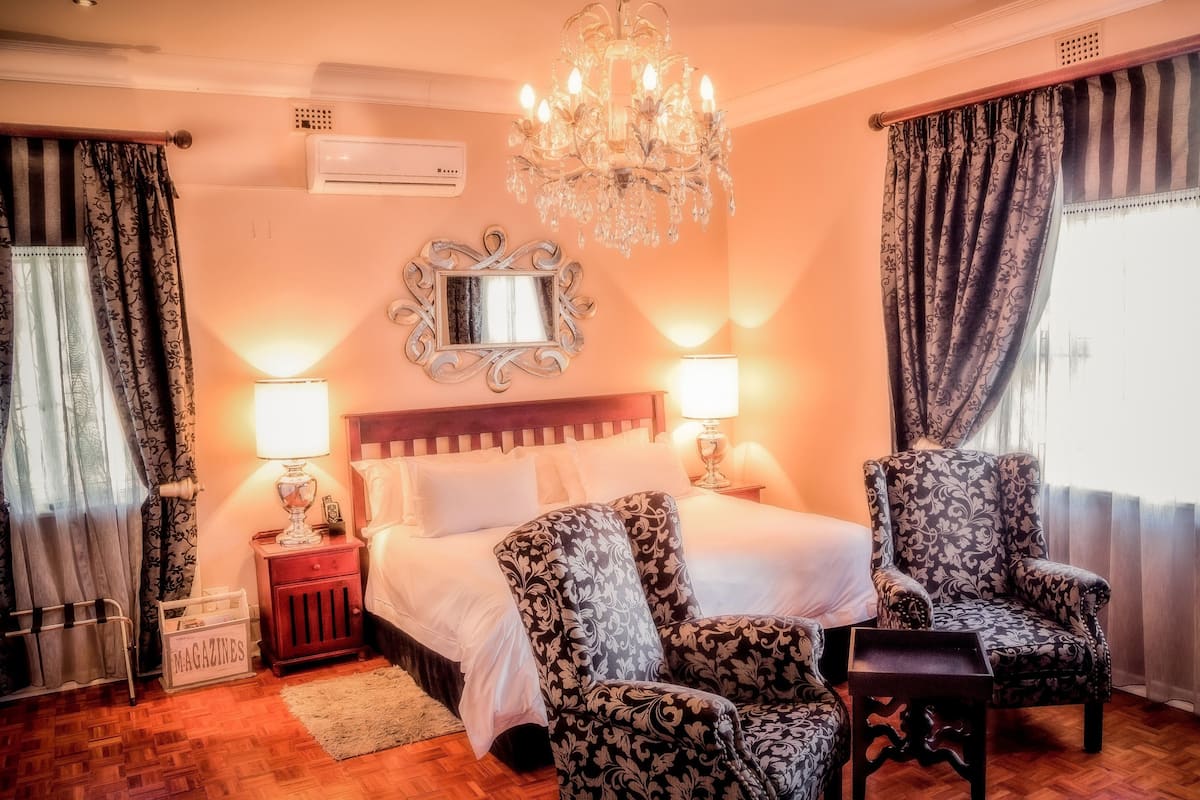 A hotel room with a large bed, two chairs, a chandelier, and a mirror on the wall.