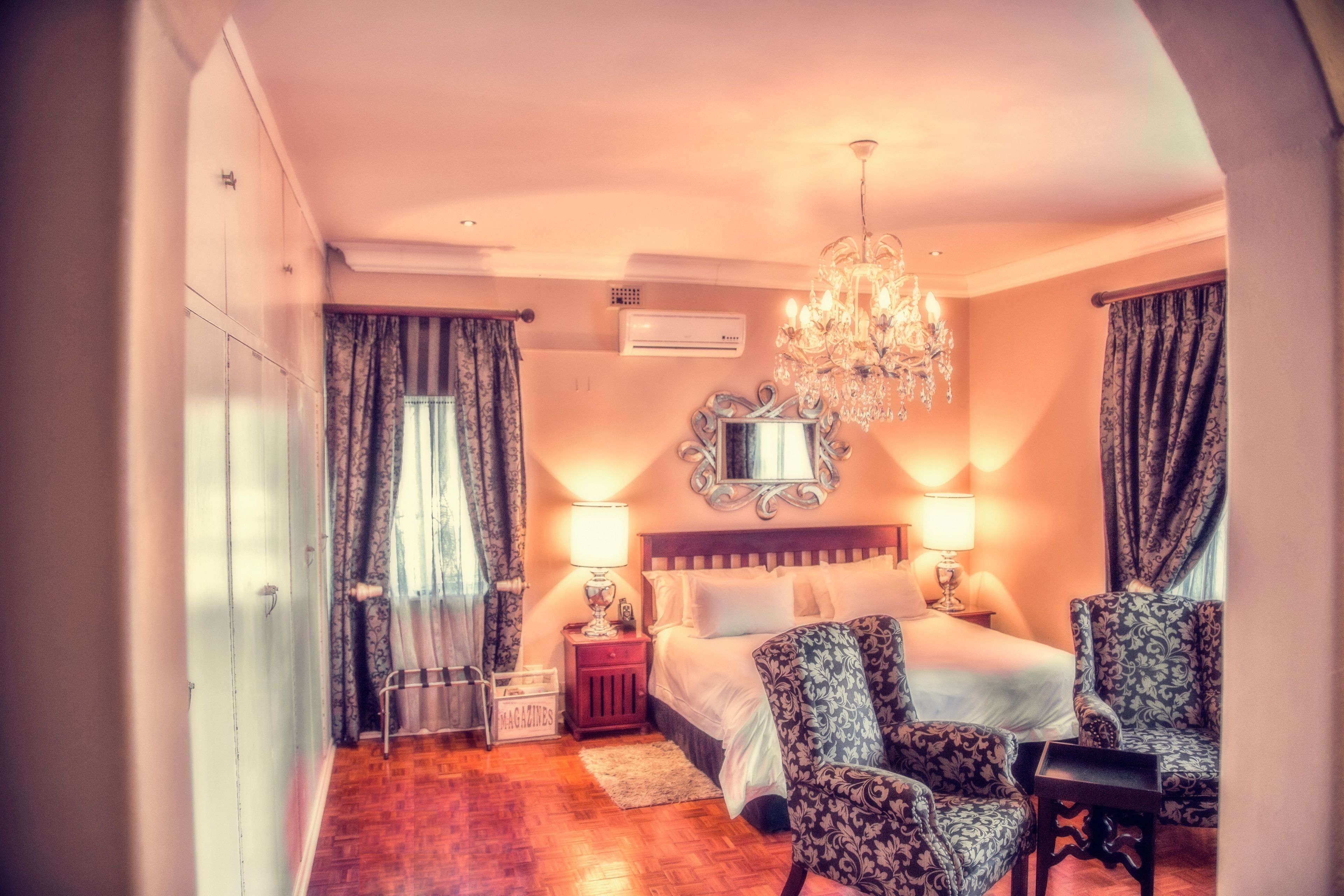 A hotel room with a bed, two chairs, a chandelier, a mirror, and an air conditioning unit.