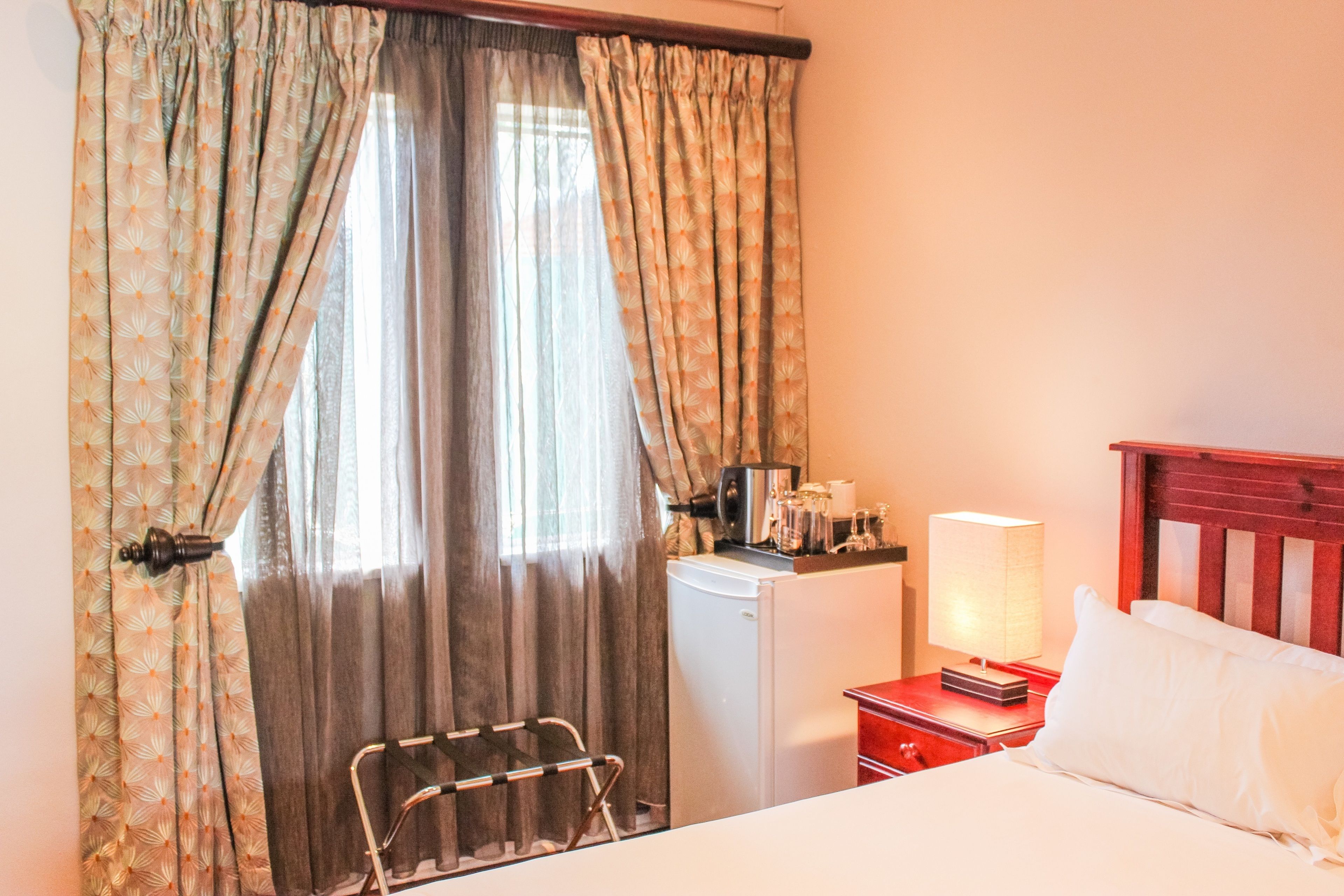 A hotel room with a bed, a desk with a lamp, a small refrigerator, and curtains.