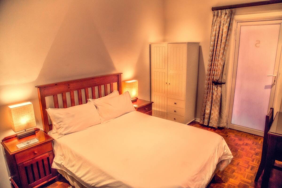 A neatly made bed in a room with a wooden headboard, a nightstand with a lamp, a wardrobe, and a door with a curtain.