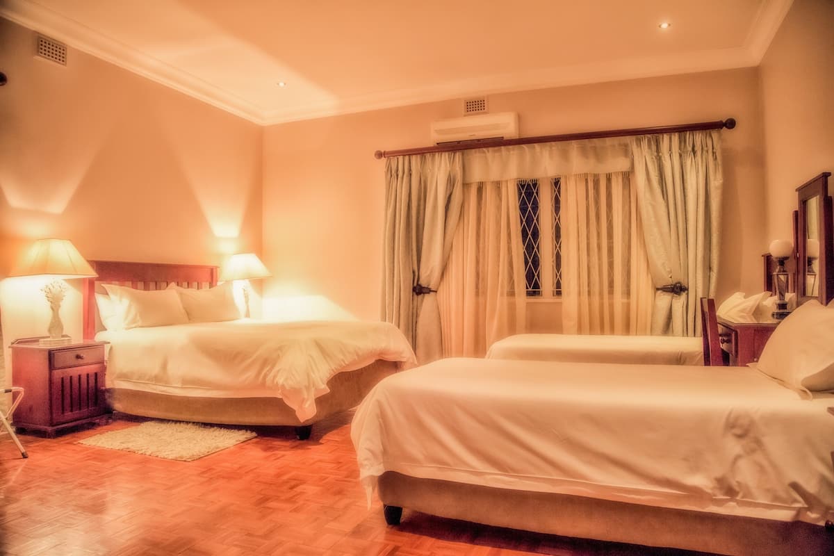A hotel room with two beds, a wooden floor, a window with curtains, and a nightstand with a lamp.