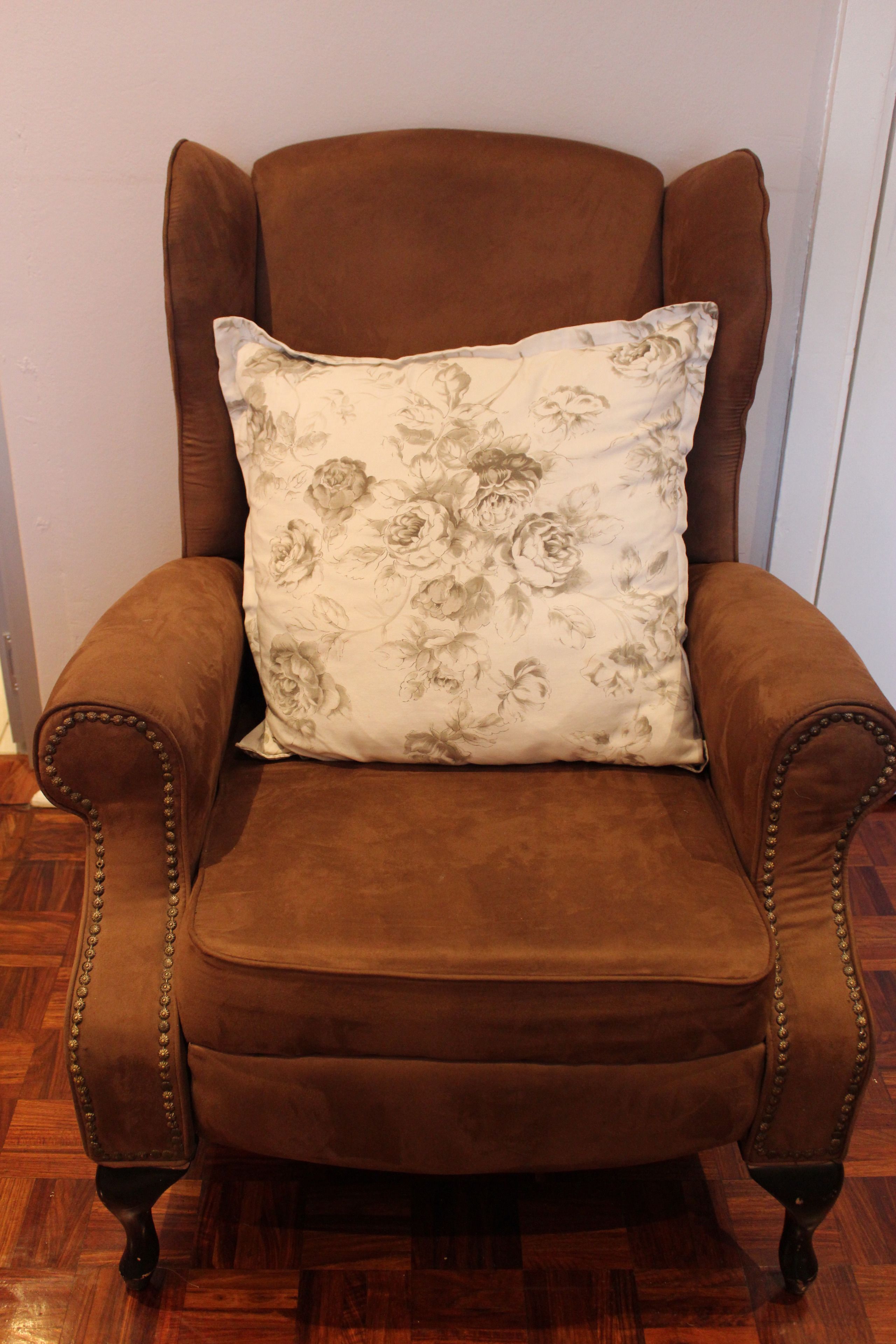A brown armchair with a floral pillow, positioned against a white wall on a wooden floor.
