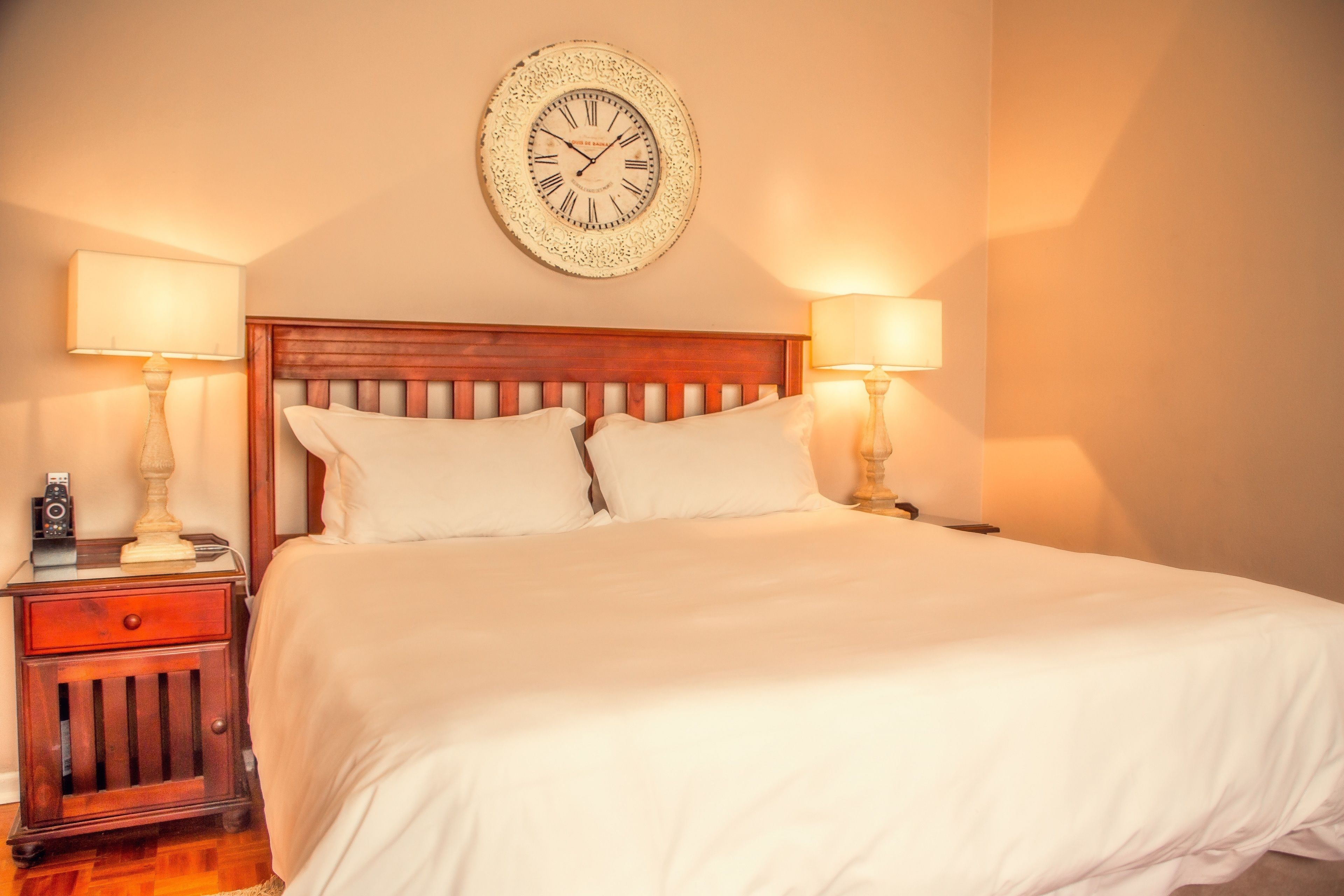 Una cama bien hecha con cabecera de madera, dos mesitas de noche con lámparas y un reloj de pared.