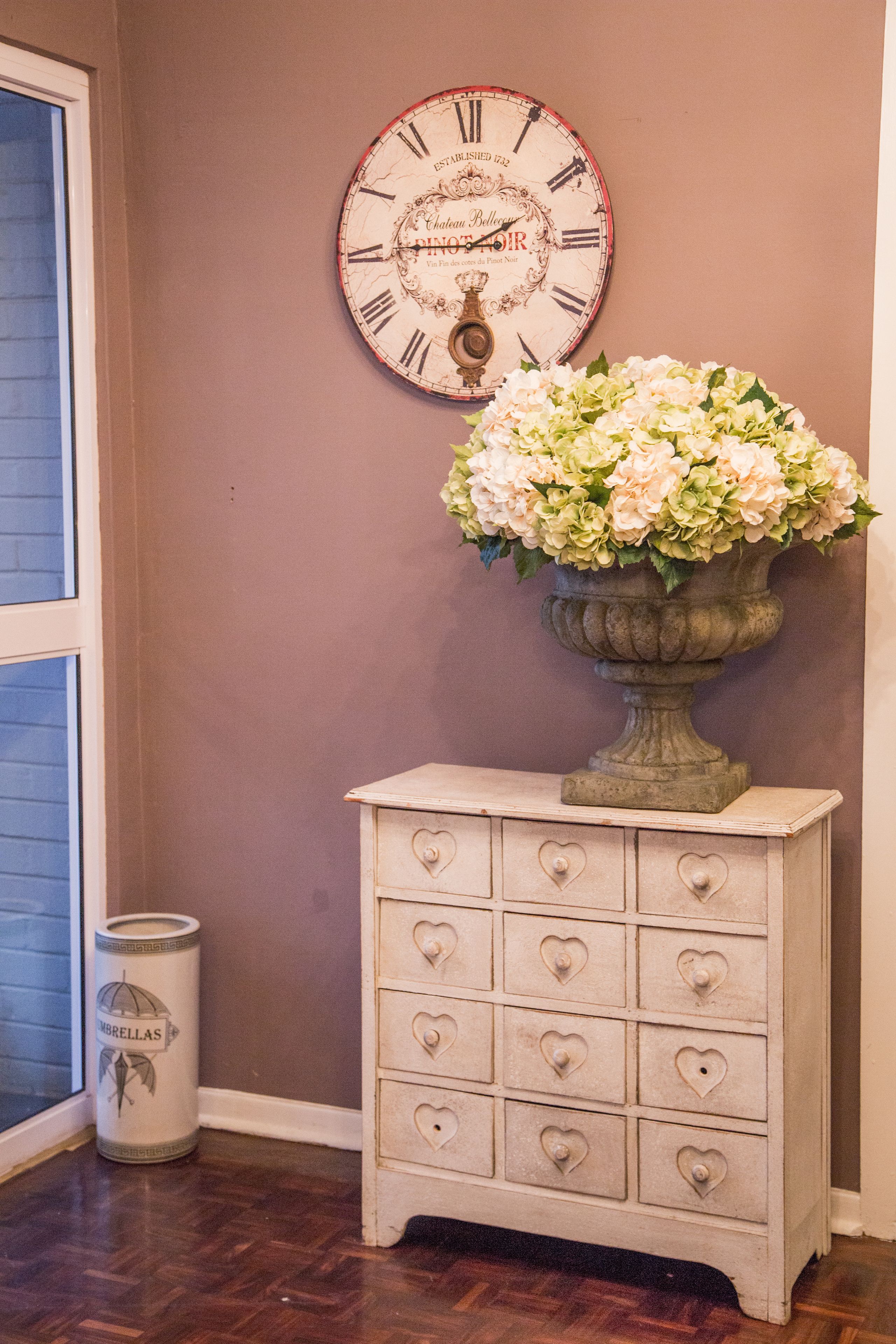 A room with a clock, a vase of flowers, and a chest of drawers.