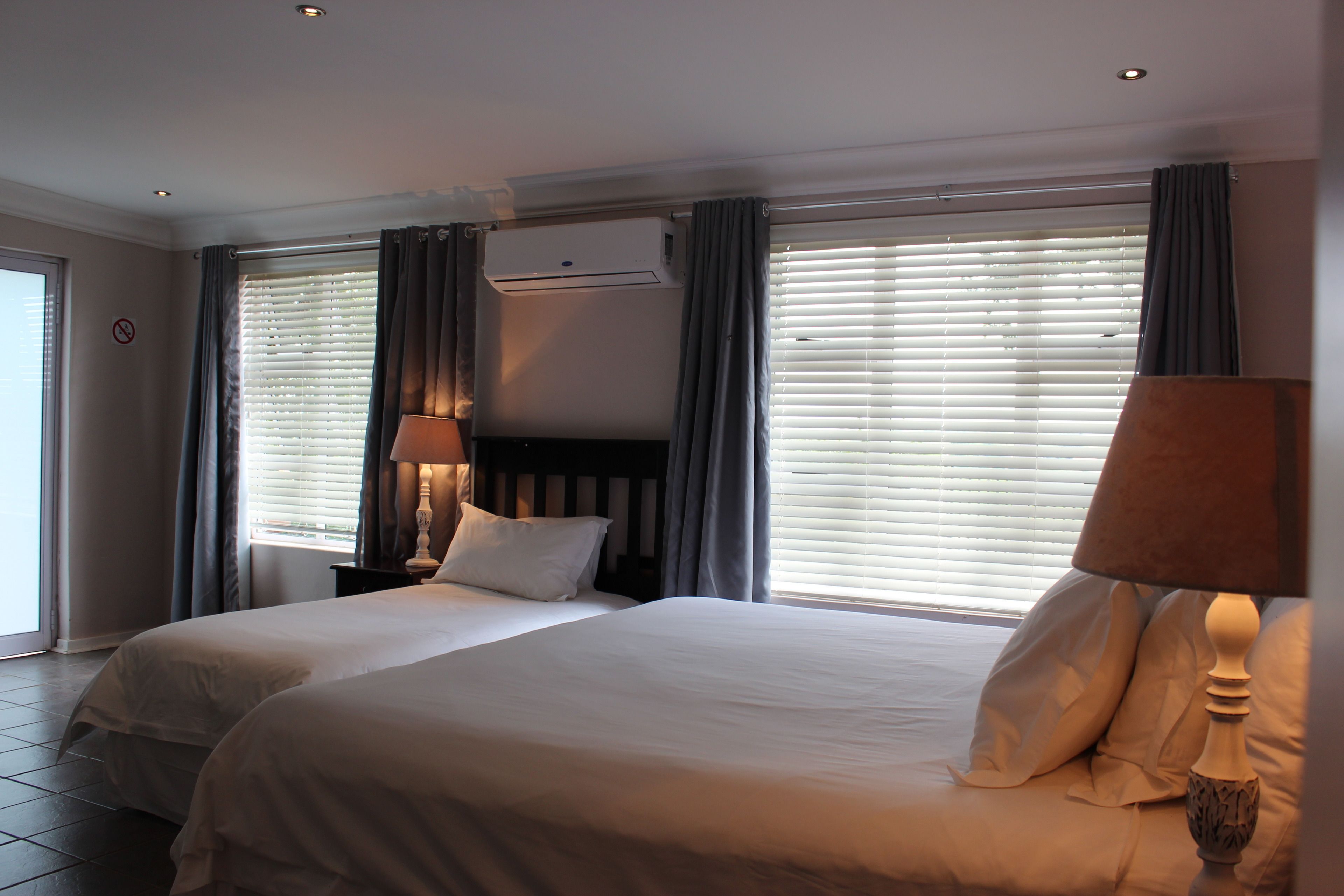 A hotel room with two beds, a window with blinds, a wall-mounted air conditioner, and a lamp on the nightstand.