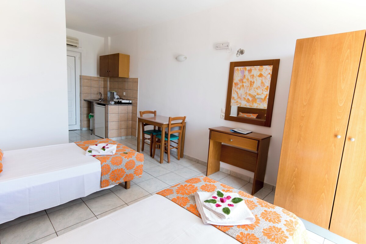 A hotel room with two beds, a desk, a chair, a wardrobe, a small kitchenette, and a bathroom.