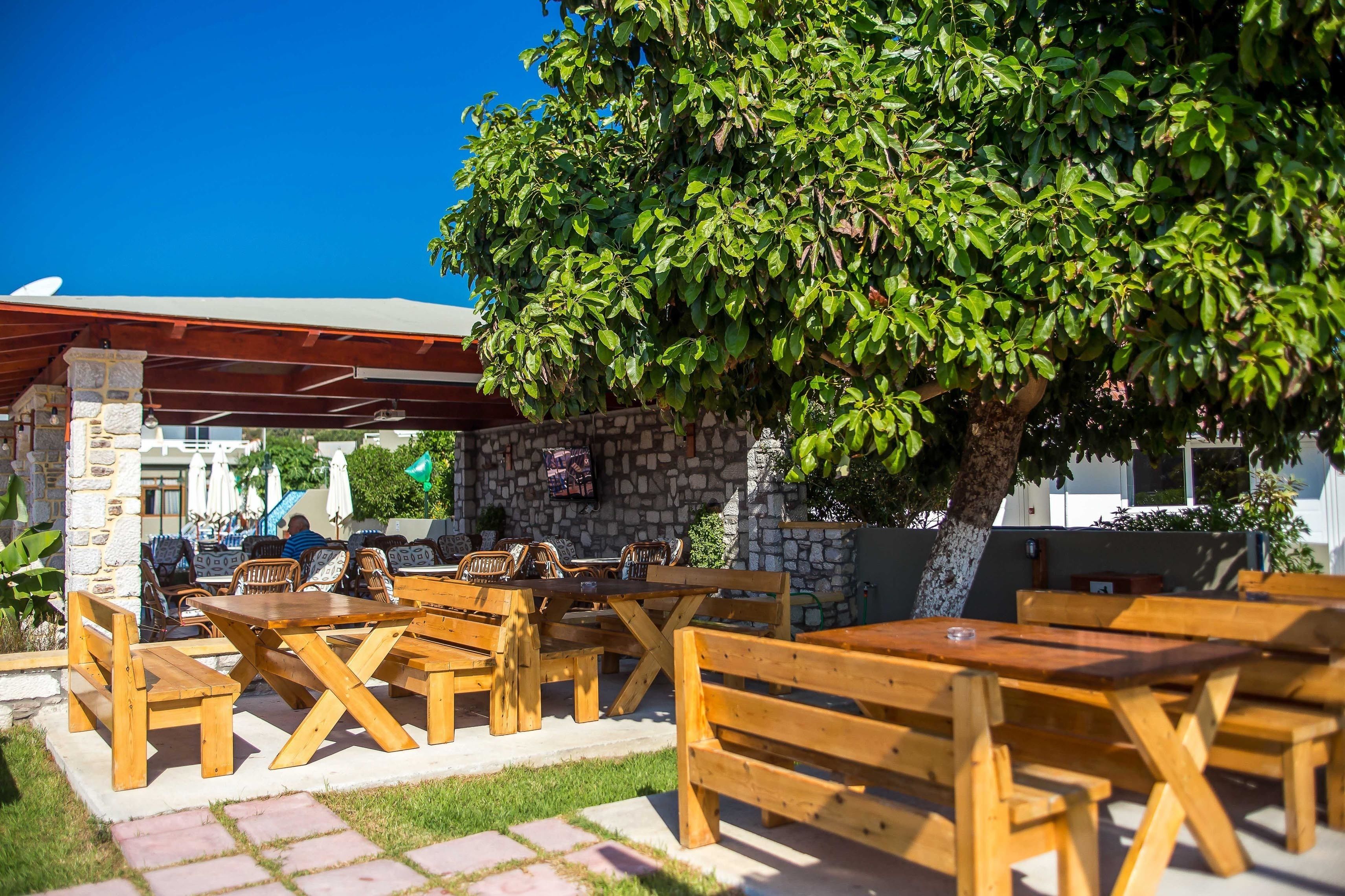 An outdoor seating area with wooden tables and benches, a stone wall, and a large tree providing shade.