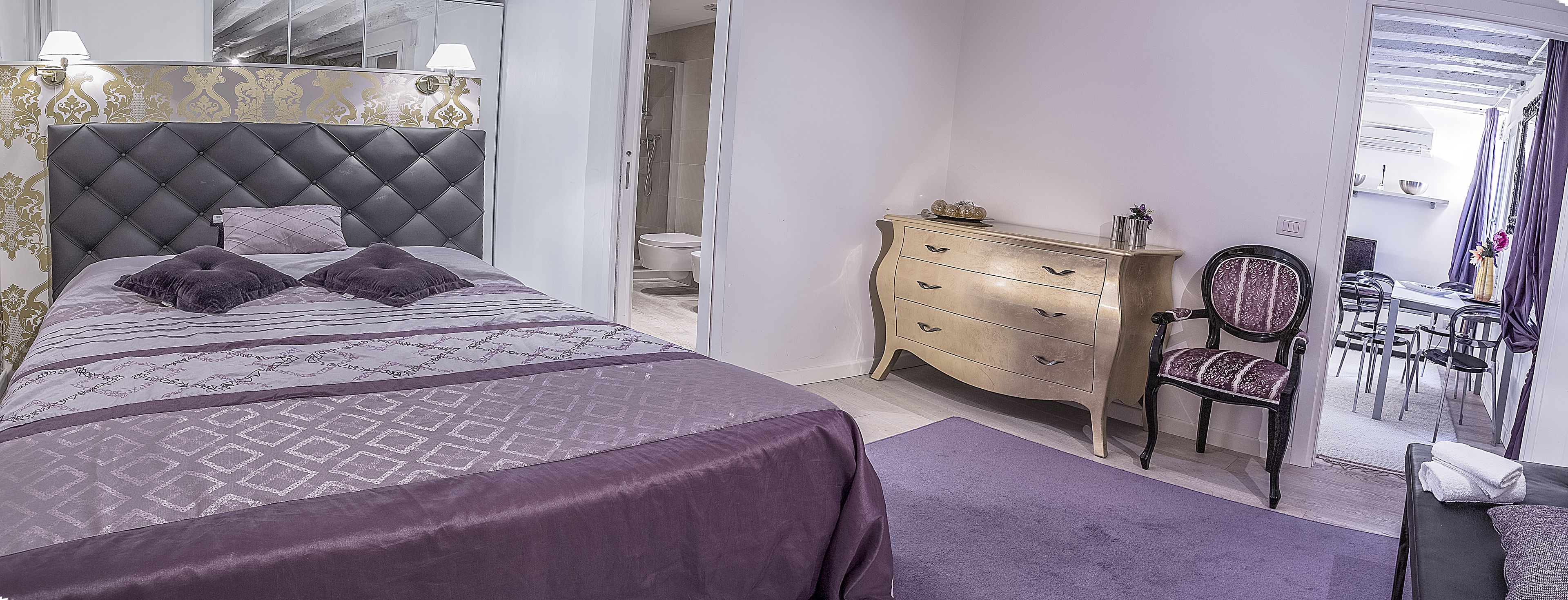 A bedroom with a tufted headboard, a bed with patterned bedding, a dresser, a chair, and a small table with a vase.